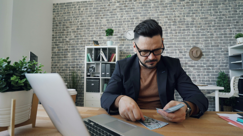 A man is at a desk with a laptop and cash in front of him. He's counting the cash he saved from Simplicity Financial's accounting and outsourcing services.