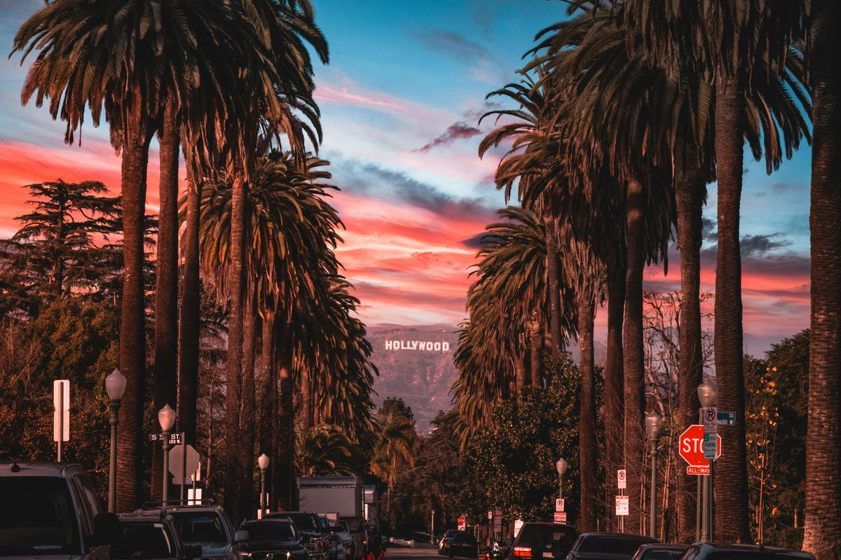 A sunset is glowing pink behind a palm tree lined street in Los Angeles. The Hollywood sign is visible in the far distance right in the middle of the view.