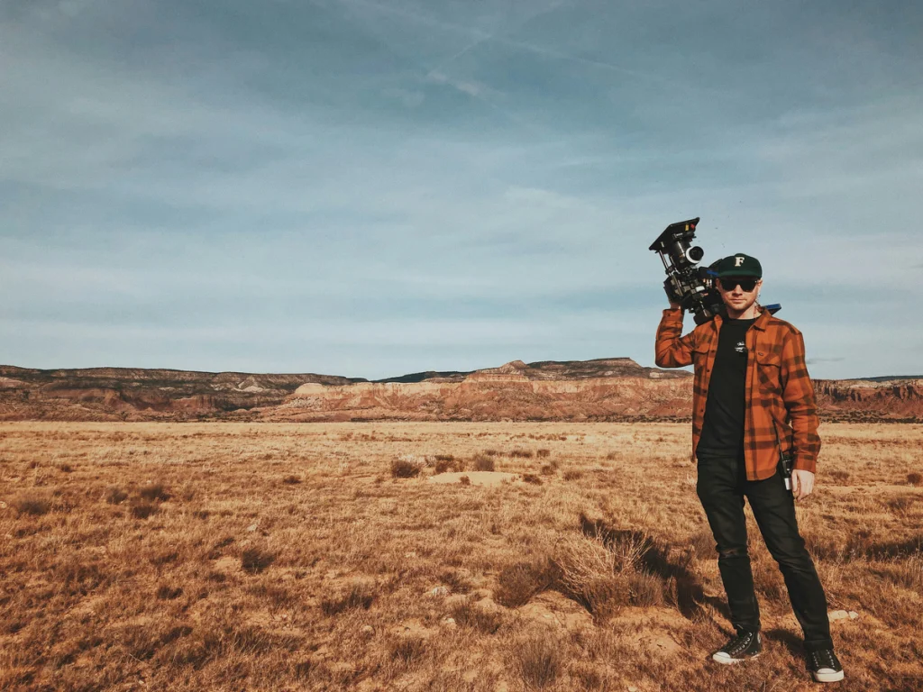 A camera operator is standing in a field with a handheld camera over his shoulder. He needs an entertainment industry CPA to support his project.