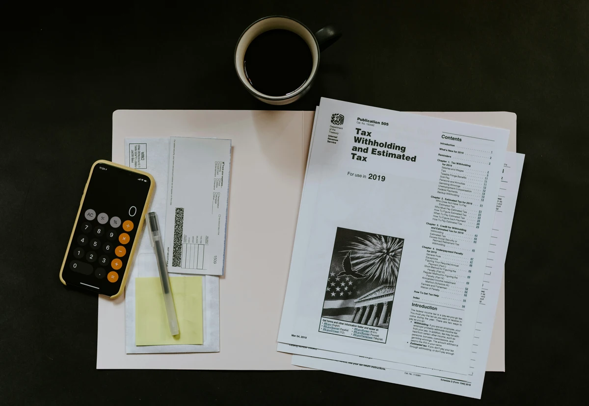 A pile of documents, a calculator, pens, and a cup of coffee are on a dark desk. Someone is learning how to read tax returns.