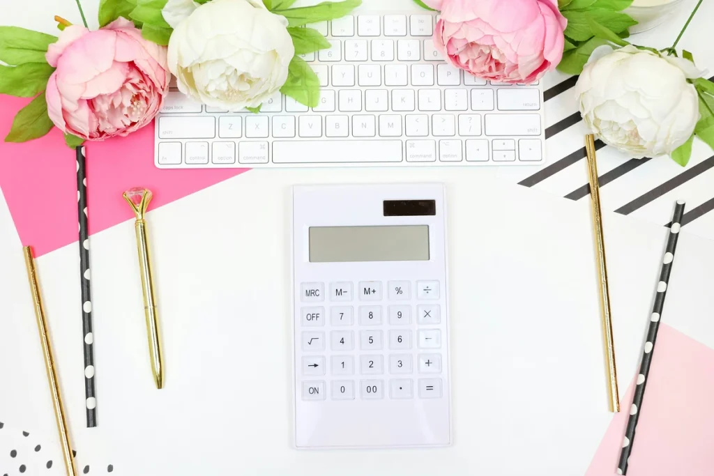 A white calculator on a white desk, also with a white keyboard, pink envelope, pink and white flowers, and various stationary like pencils. This is what you need in front of you when searching 'how do I report a 1099-s on my tax return'.